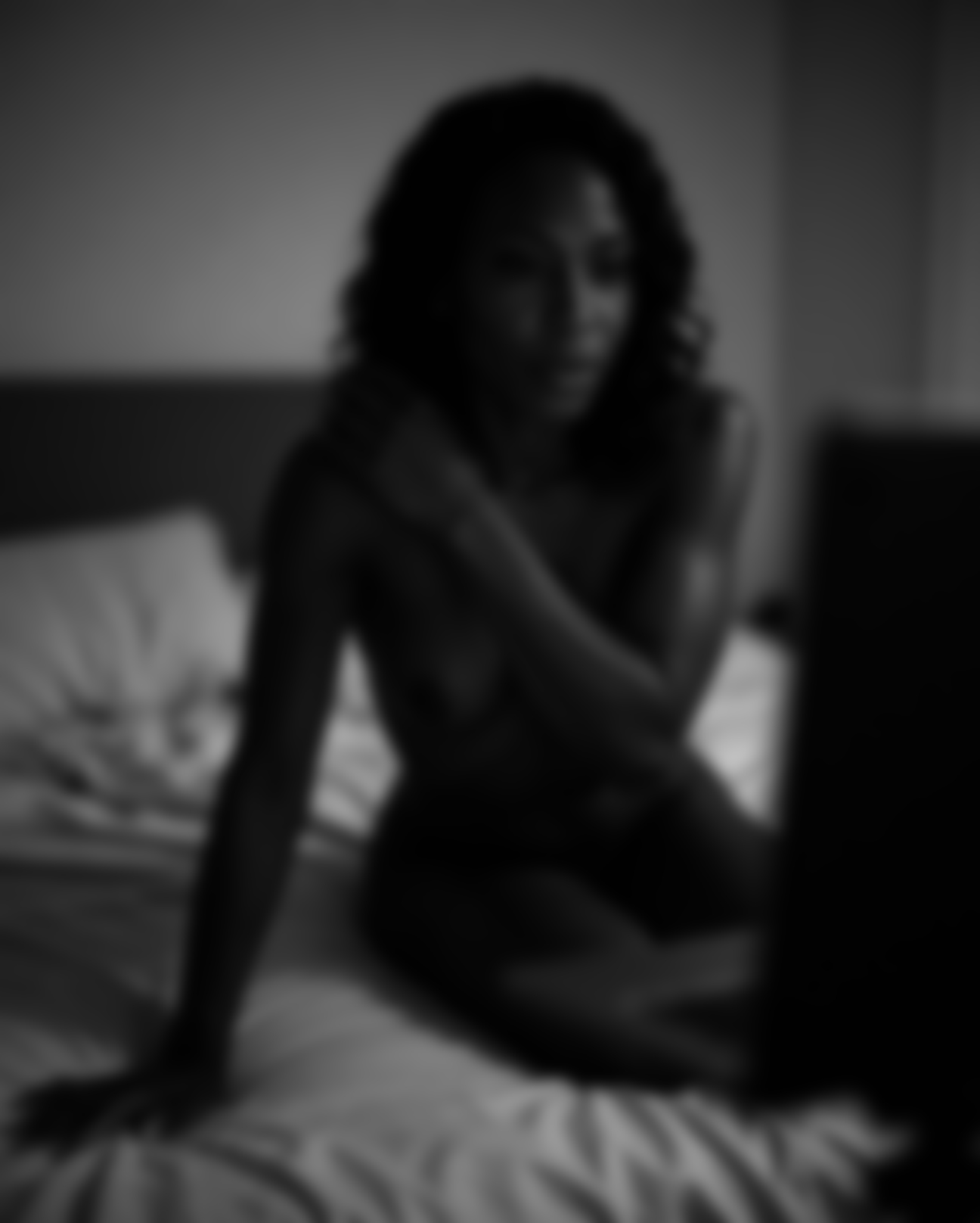 Soft-focus monochrome photograph of a woman seated on a bed.