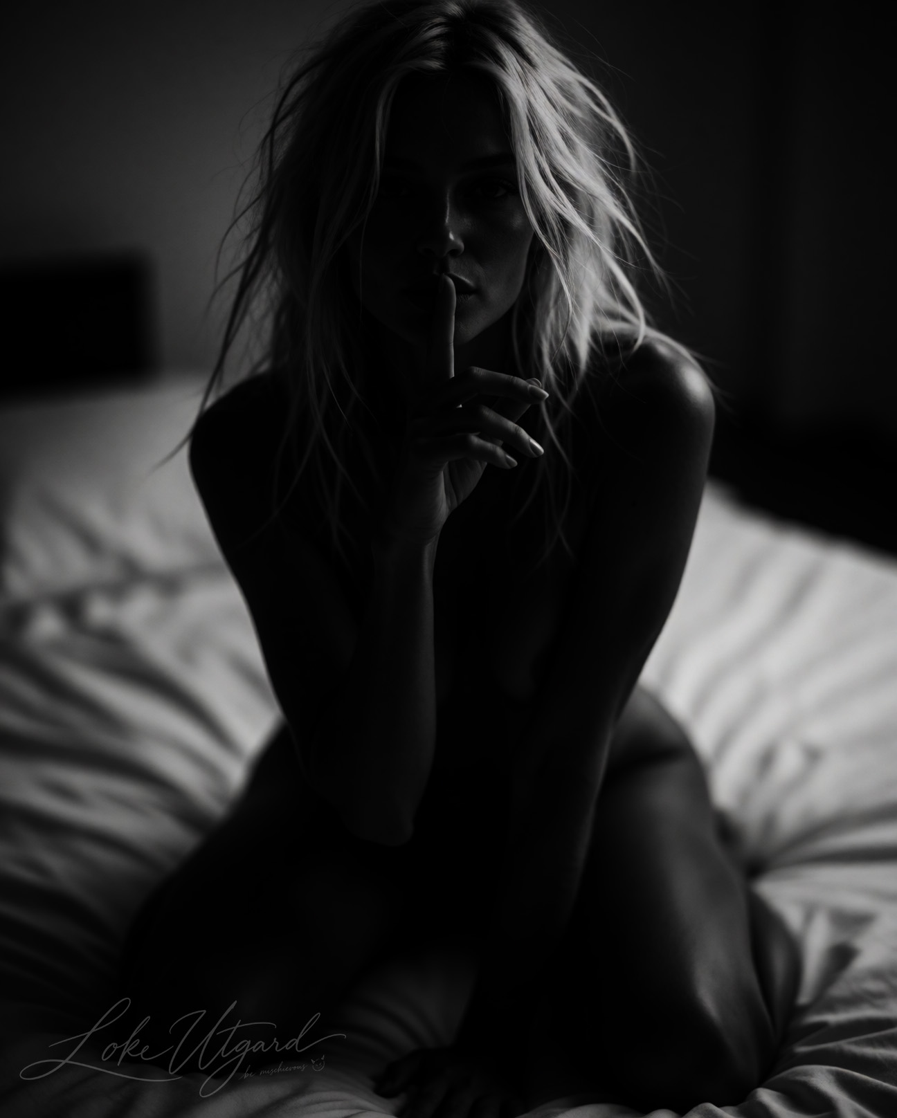 Low-key monochrome portrait of a woman on a bed with a finger to her lips.