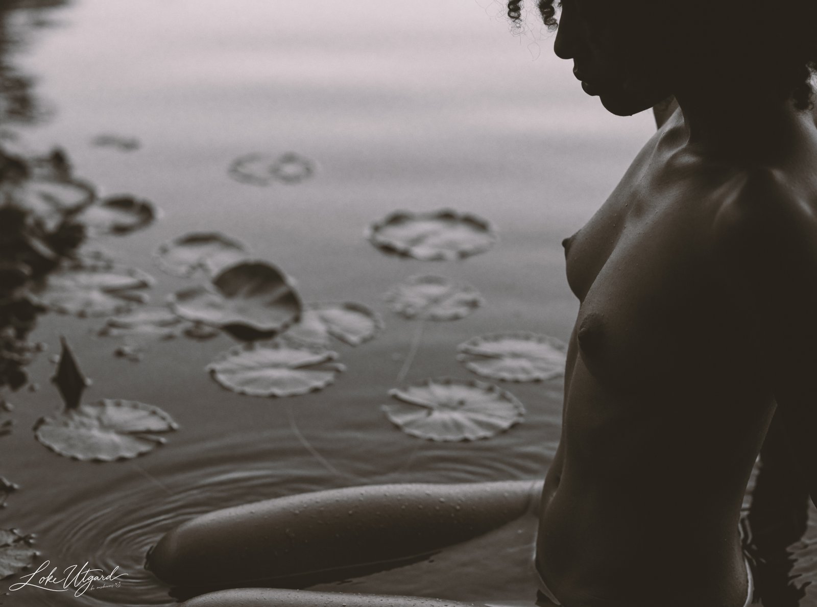 Side-profile monochrome portrait of Tania seated nude in a lake among lily pads